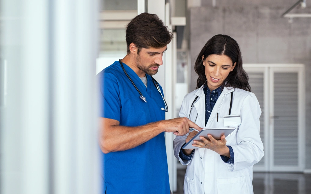 Brunette doctor wearing a lab coat holding an iPad. Next to her, a medical professional wearing blue scrubs is pointing at the iPad while they discuss.