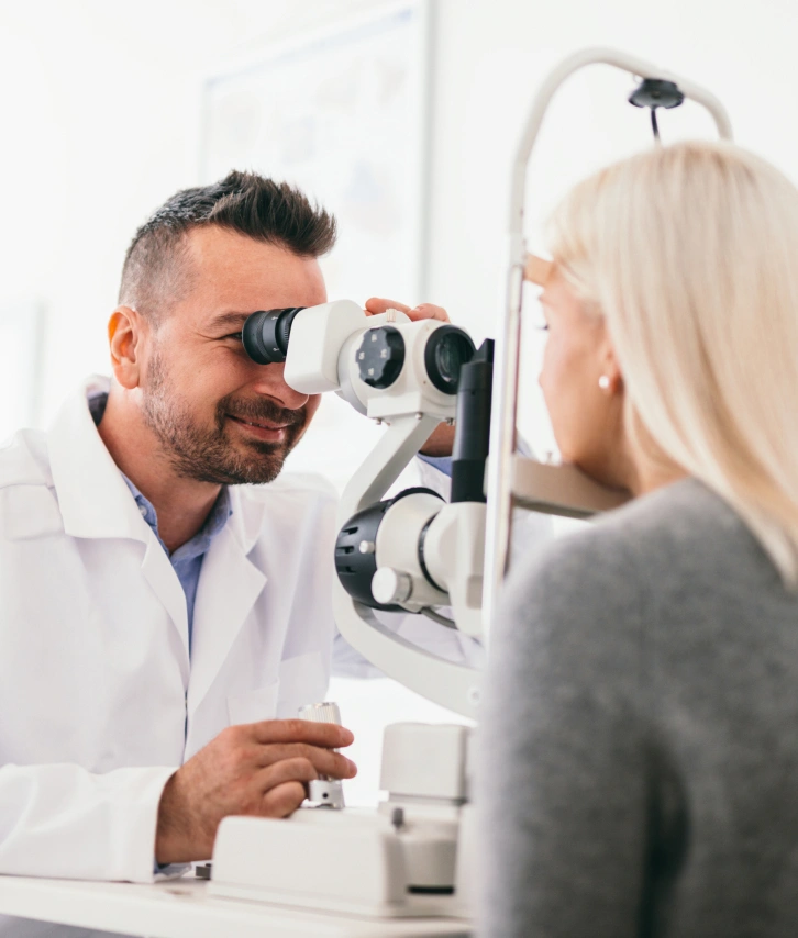 An eye doctor wearing a lab coat looking through a lens at his patient's eyes. The patient is a blonde woman with her back to the camera.