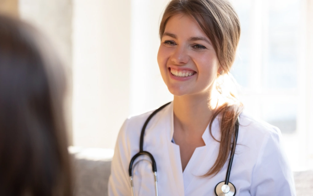 Smiling brunette doctor with her hair in a low ponytail. She's wearing a lab coat and has a stethoscope draped around her neck.